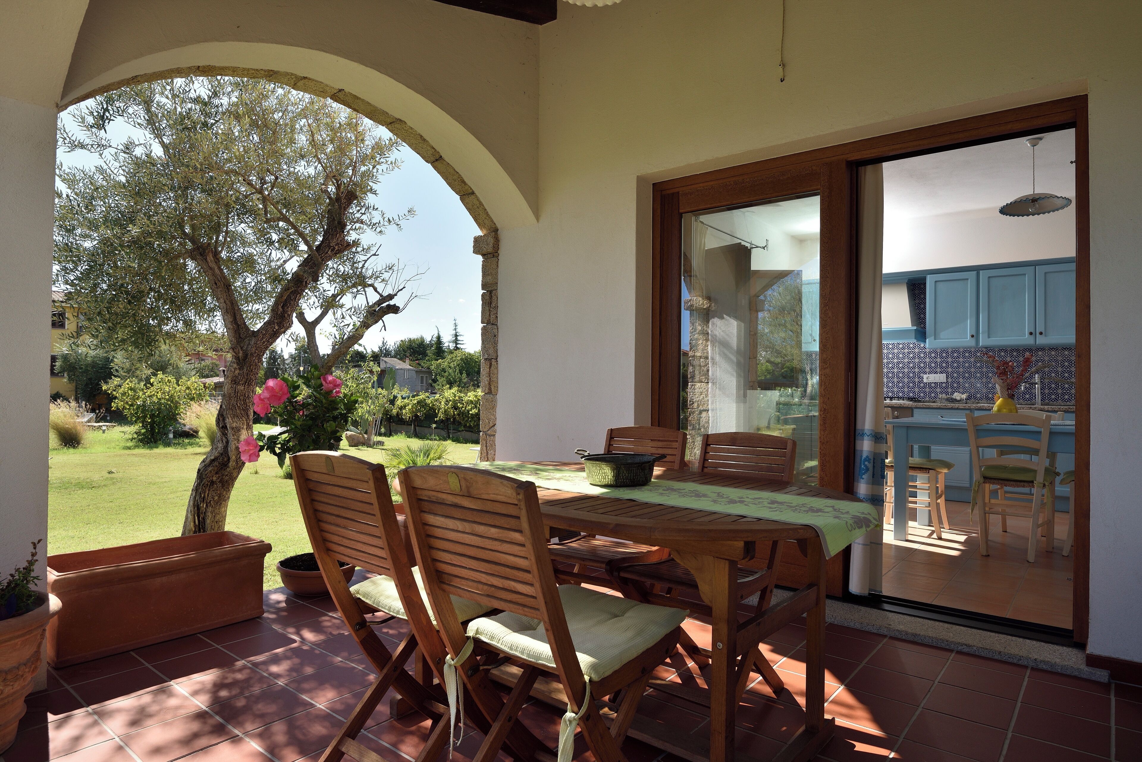 Eine Terrasse im Freien mit Tisch und Stühlen, einem Blumentopf und Blick auf eine Küche durch eine Glastür.