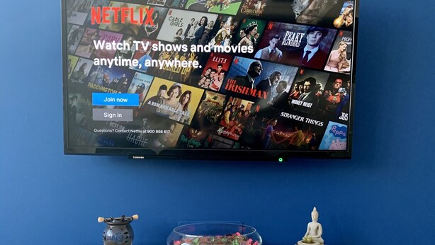 Un televisor de pantalla plana montado en una pared azul que muestra la interfaz de Netflix con diversas series y películas. Debajo del televisor, hay un mueble blanco con elementos decorativos, incluyendo una estatua de Buda, una planta pequeña y un cuenco decorativo con flores.