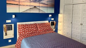 Un dormitorio con cama, un cuadro en la pared que muestra un atardecer sobre el agua, un armario y una mesita de noche con lámpara.