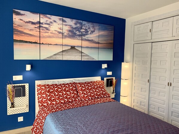 A bedroom with a bed, a wall-mounted artwork of a sunset over water, a wardrobe, and a bedside table with a lamp.