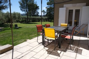 An outdoor patio with a table and chairs, a covered area, and a view of a grassy garden and trees.