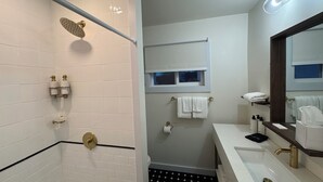 A bathroom with white subway tiles, a black and white checkered floor, a shower with a gold showerhead, a white sink with a gold faucet, and a mirror with a wooden frame.