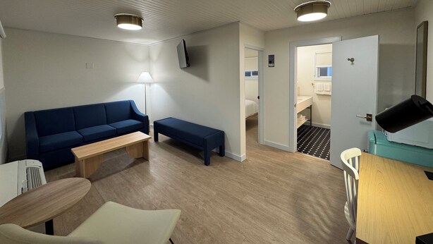 A compact room with a blue sofa, a wooden bench, a small table, and a white chair. A lamp is on the floor, and a door leads to a bathroom with a sink and a mirror.