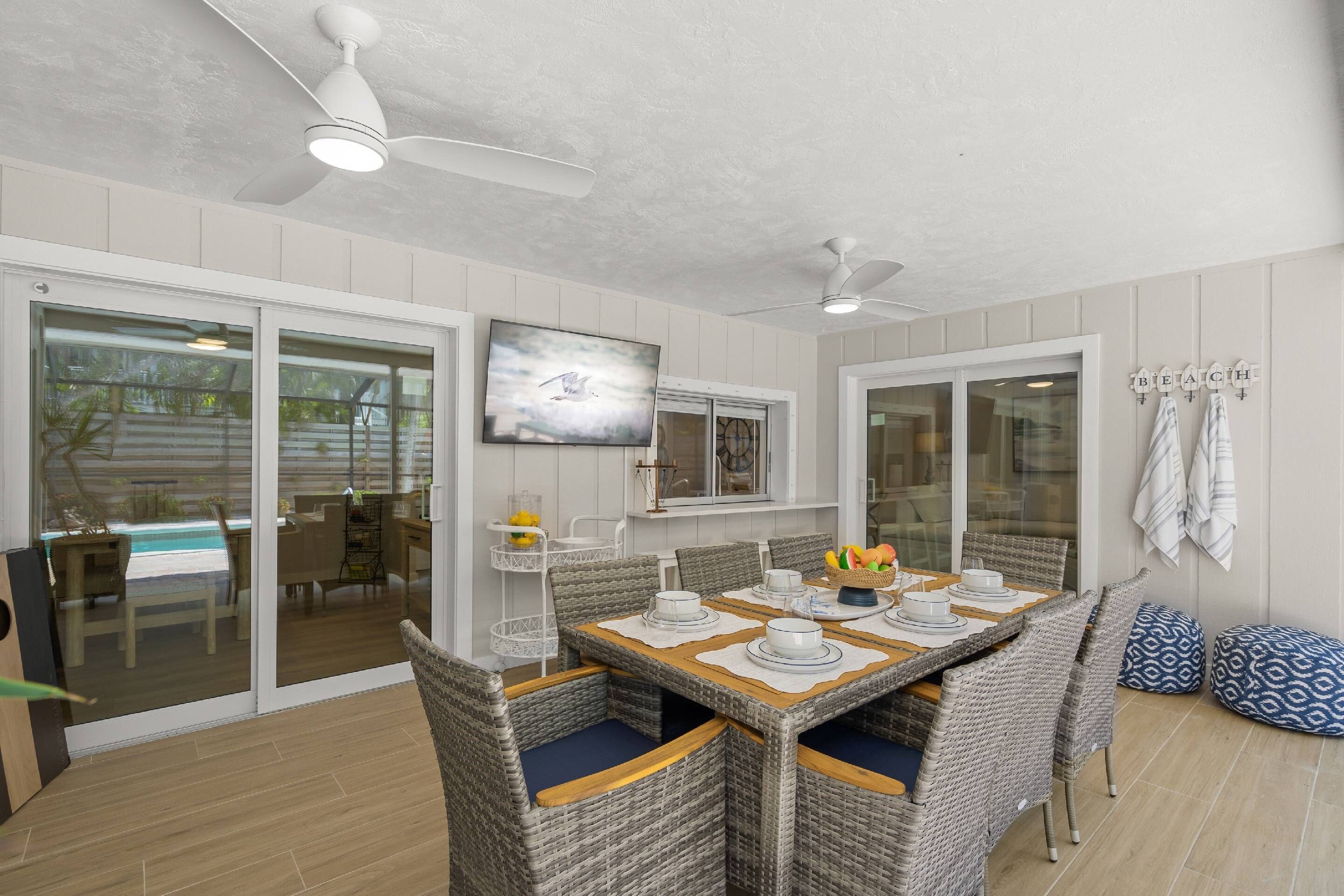 A dining area with a table set for four, surrounded by wicker chairs with blue cushions. There are ceiling fans above and a large window with a view of the outdoors.