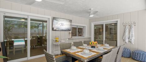 A dining area with a table set for four, surrounded by wicker chairs with blue cushions. There are ceiling fans above and a large window with a view of the outdoors.