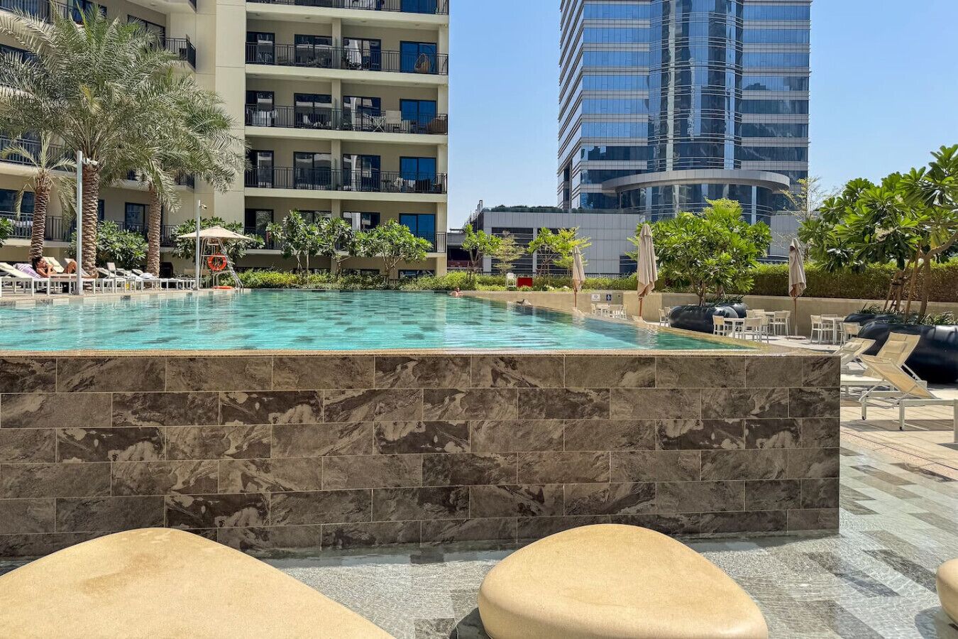 A swimming pool with lounge chairs and umbrellas, surrounded by palm trees and a modern building in the background.