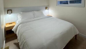 A bedroom with a bed, a nightstand, a wall-mounted lamp, and a window with blinds. - King Motel Room With Balcony - NKB (Geneva)