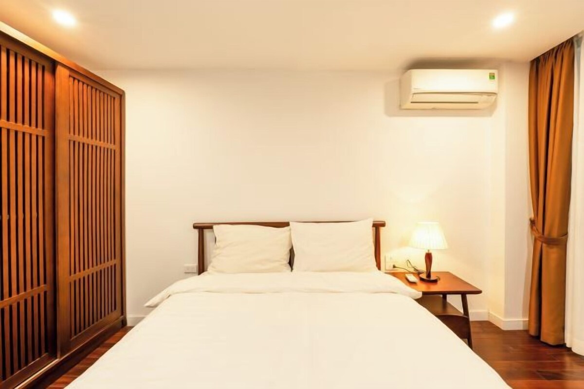 A hotel room with a bed, a nightstand with a lamp, an air conditioning unit, and a wooden wardrobe.