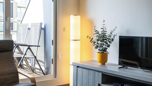 Una sala de estar moderna con un sofá, un televisor sobre un soporte de madera, una planta en maceta y un ventanal con persianas.