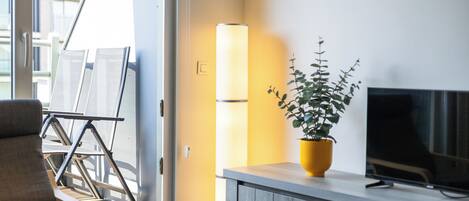 Een moderne woonkamer met een bank, een televisie op een houten dressoir, een potplant en een groot raam met jaloezieën.