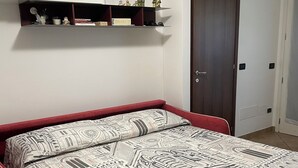 Une chambre à coucher avec un lit, un banc recouvert d’un tissu rouge, un mur blanc, une porte en bois et une étagère ornée d’objets décoratifs.
