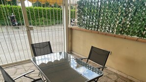 Une terrasse extérieure dotée d’une table à plateau en verre et de quatre chaises, entourée d’un sol en pierre et d’une clôture en métal.
