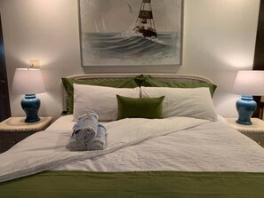 Una cama bien hecha con ropa de cama verde, dos lámparas azules sobre mesitas de noche y un cuadro de un velero en la pared.