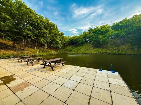 Un muelle de hormigón con mesas de picnic y bancos que da a un río tranquilo, rodeado de abundante vegetación.