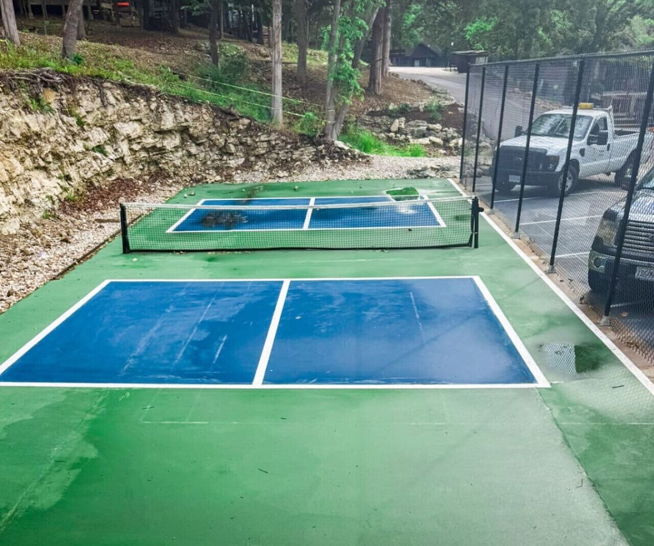 Ein Tennisplatz im Freien mit Netz, umgeben von einem Zaun und einem Parkplatz mit Fahrzeugen.