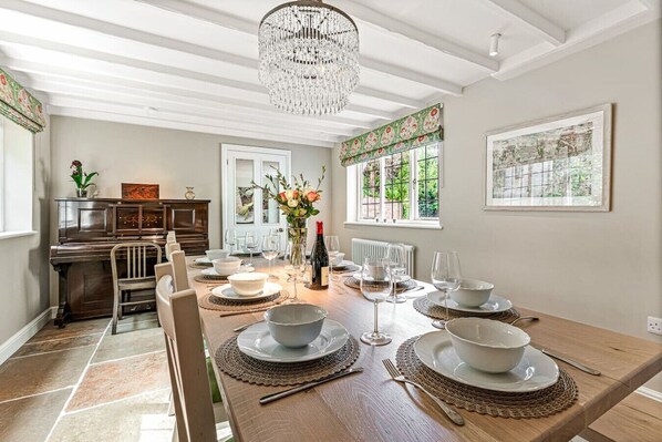A dining room with a long wooden table set for six, surrounded by chairs. A chandelier hangs from the ceiling, and there is a piano in the background.