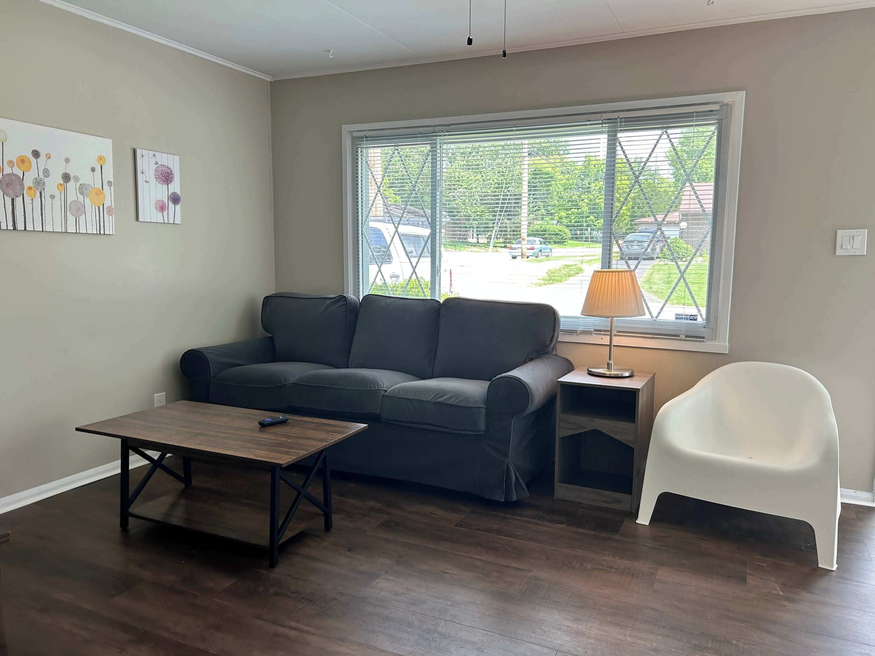 Un salon avec un canapé gris foncé, une table basse en bois, une chaise blanche et une fenêtre avec des stores.