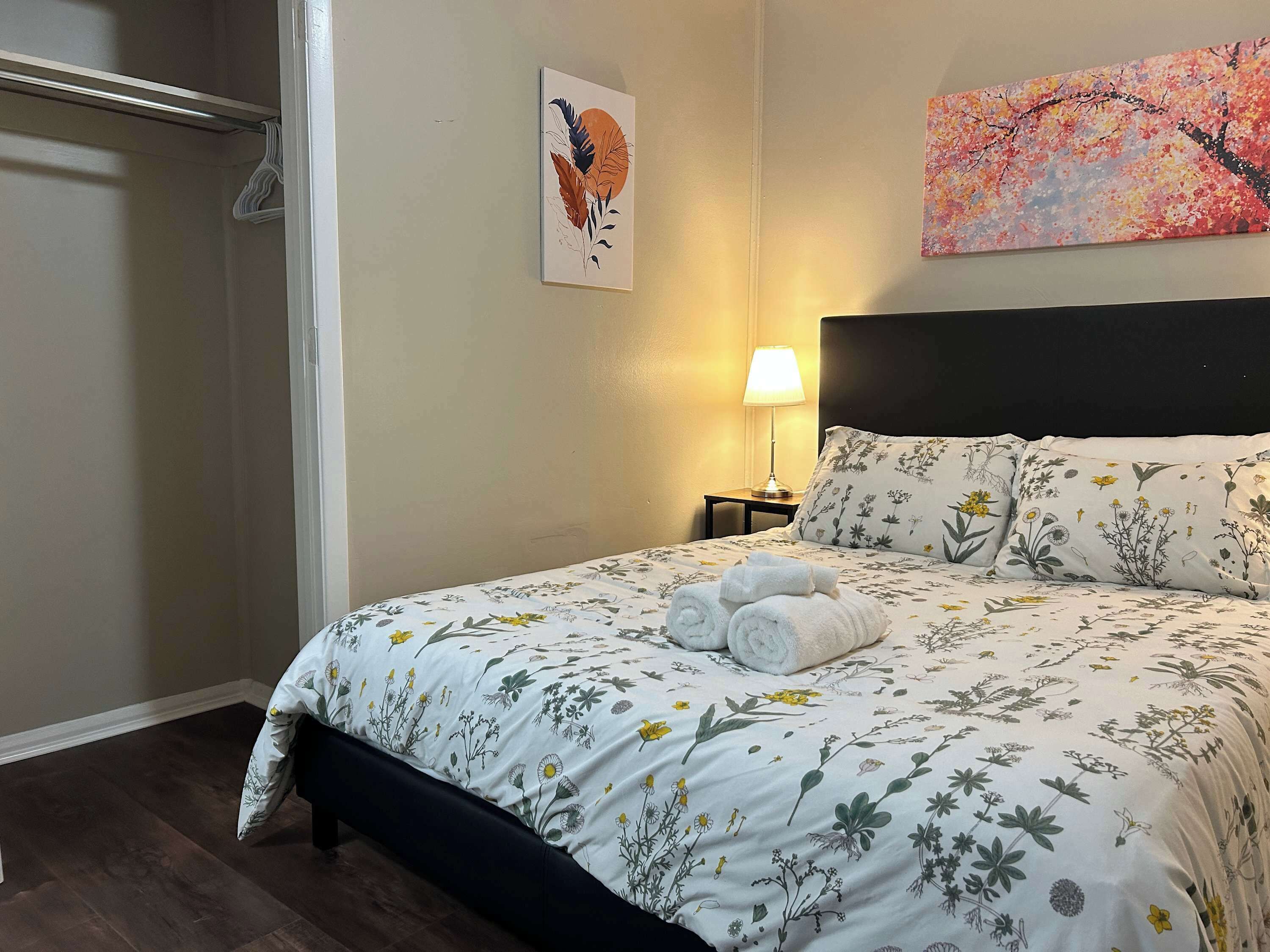 A hotel room with a floral bedspread, a headboard, a nightstand with a lamp, and a painting on the wall.