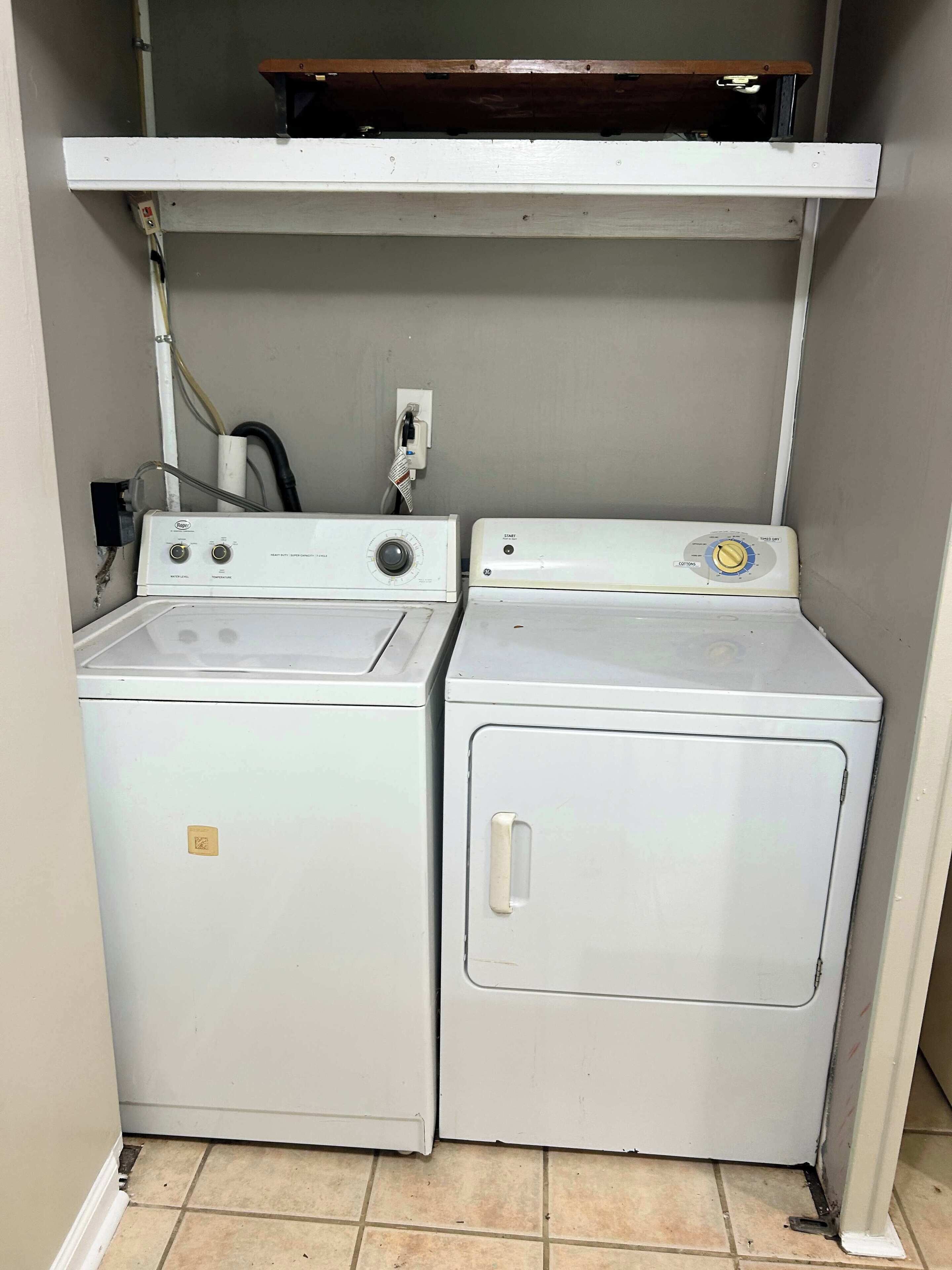 A compact laundry area with two white washing machines and a dryer, equipped with a wooden shelf above.
