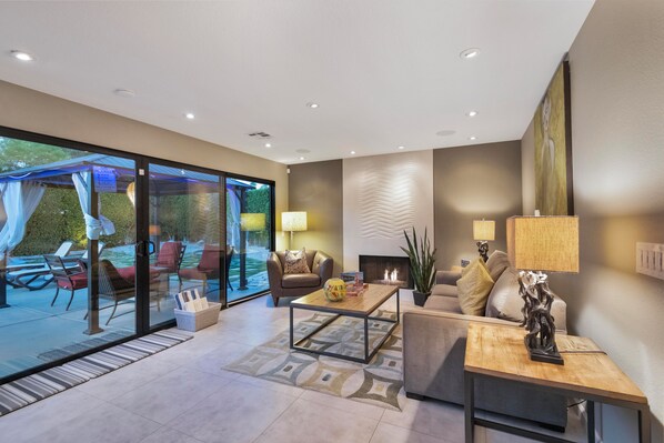 A modern living room with a glass door leading to an outdoor seating area, a fireplace, and a coffee table with decorative items.