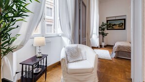 A room with a white chair, a small table with a lamp, a bed, and a painting on the wall. - Fabulous Cozy Pantheon Apartment (Roma)