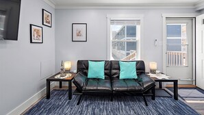 A modern living room with a black leather sofa, two teal cushions, a large window with a view of buildings, and a flat-screen TV mounted on the wall. - Two Bedroom Oceanside Condo Steps from the Beach (Ocean City)