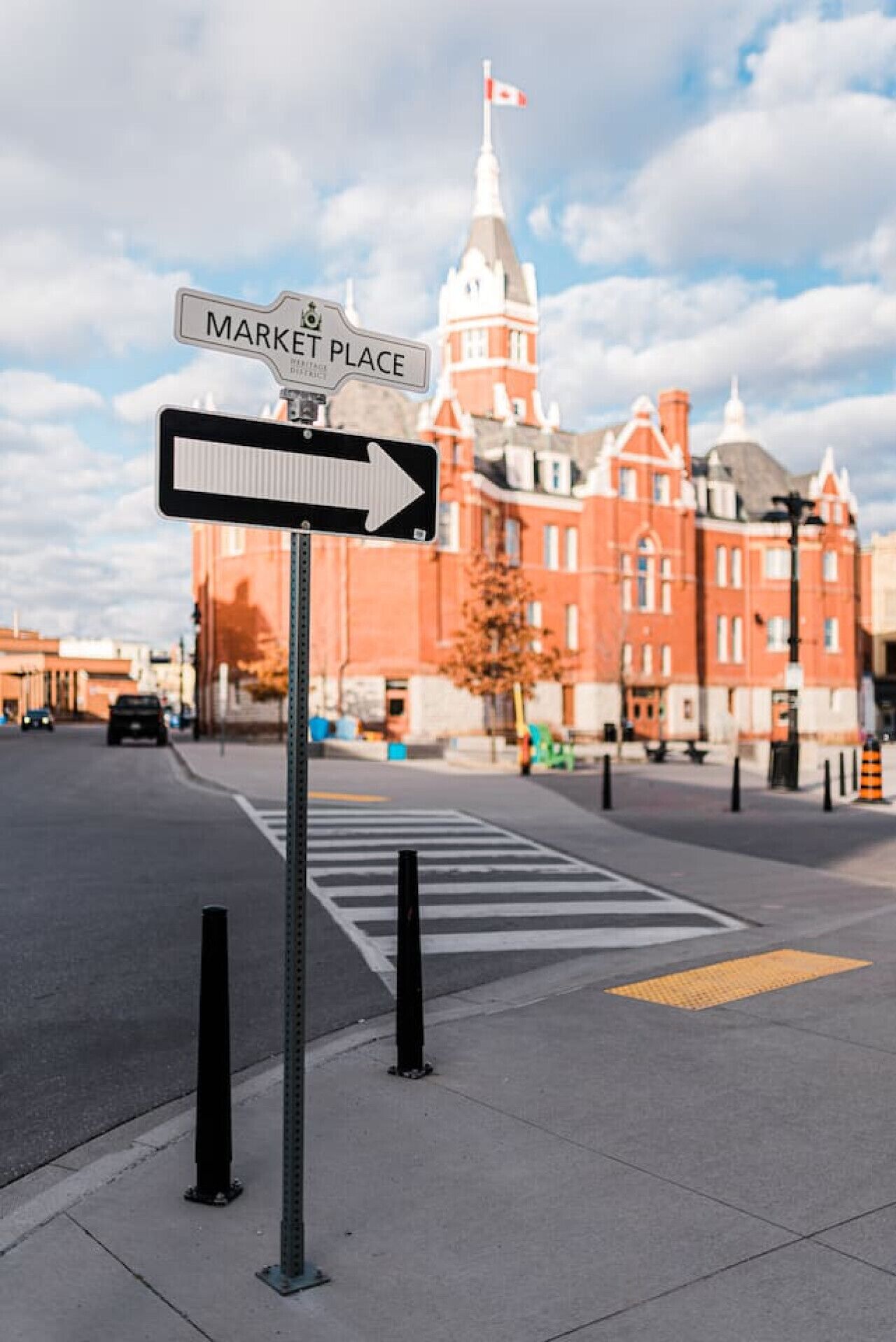 Un panneau de signalisation indiquant « PLACE DU MARCHÉ » et une flèche pointant vers la droite, devant un bâtiment en briques rouges surmonté d’un dôme blanc et d’un drapeau.