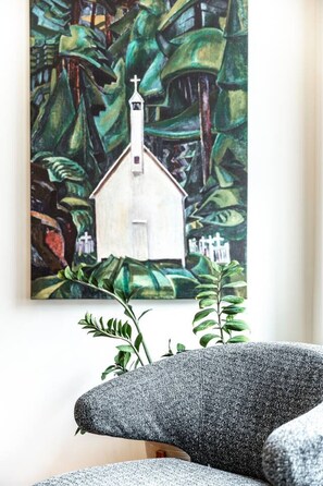 A room with a painting of a church on the wall, a grey sofa, and green plants.