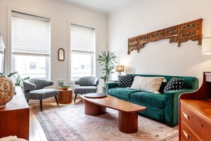 A living room with a green sofa, two armchairs, a coffee table, and a wooden cabinet. There are two windows with blinds, a potted plant, and a decorative wooden panel on the wall.