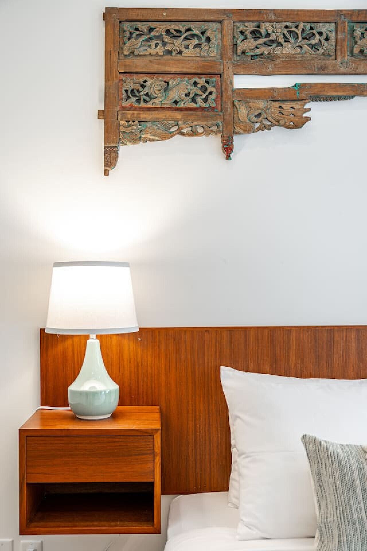 Un lit bien fait, avec un oreiller blanc et un plaid rayé, une table de chevet en bois avec une lampe bleue, et un élément décoratif en bois accroché au mur au-dessus du lit.