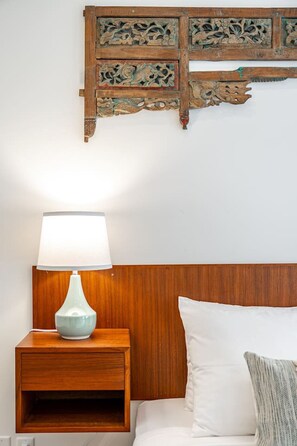 Una cama bien hecha con una almohada blanca y un plaid a rayas, una mesita de noche de madera con una lámpara azul, y un adorno de pared de madera sobre la cama.