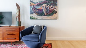 A living room with a blue armchair, a wooden cabinet, a flat-screen TV, and a colorful painting on the wall.
