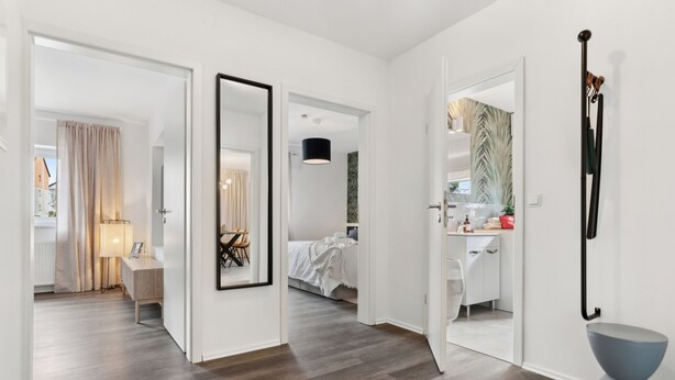 A modern hotel room with a wooden floor, a large mirror, a bed, a desk, and a bathroom visible through an open door.
