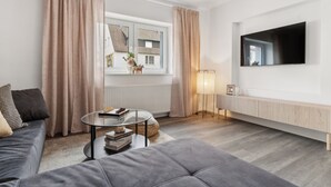 A modern living room with a grey sofa, a round coffee table, a TV mounted on the wall, and a window with curtains.