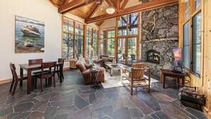 Un salón amplio con suelo de piedra, muebles de madera, ventanas grandes, una chimenea de piedra y un cuadro de un barco en la pared.