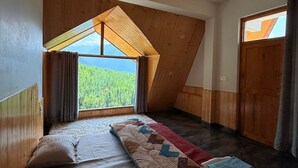 A bedroom with a sloped ceiling, a bed with a quilt, a window with curtains, and a view of greenery. - Noni’s Dream Villa (Kullu)