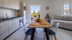 Una cocina moderna con armarios blancos, una mesa de comedor de madera con sillas transparentes, un frutero y vistas al exterior.