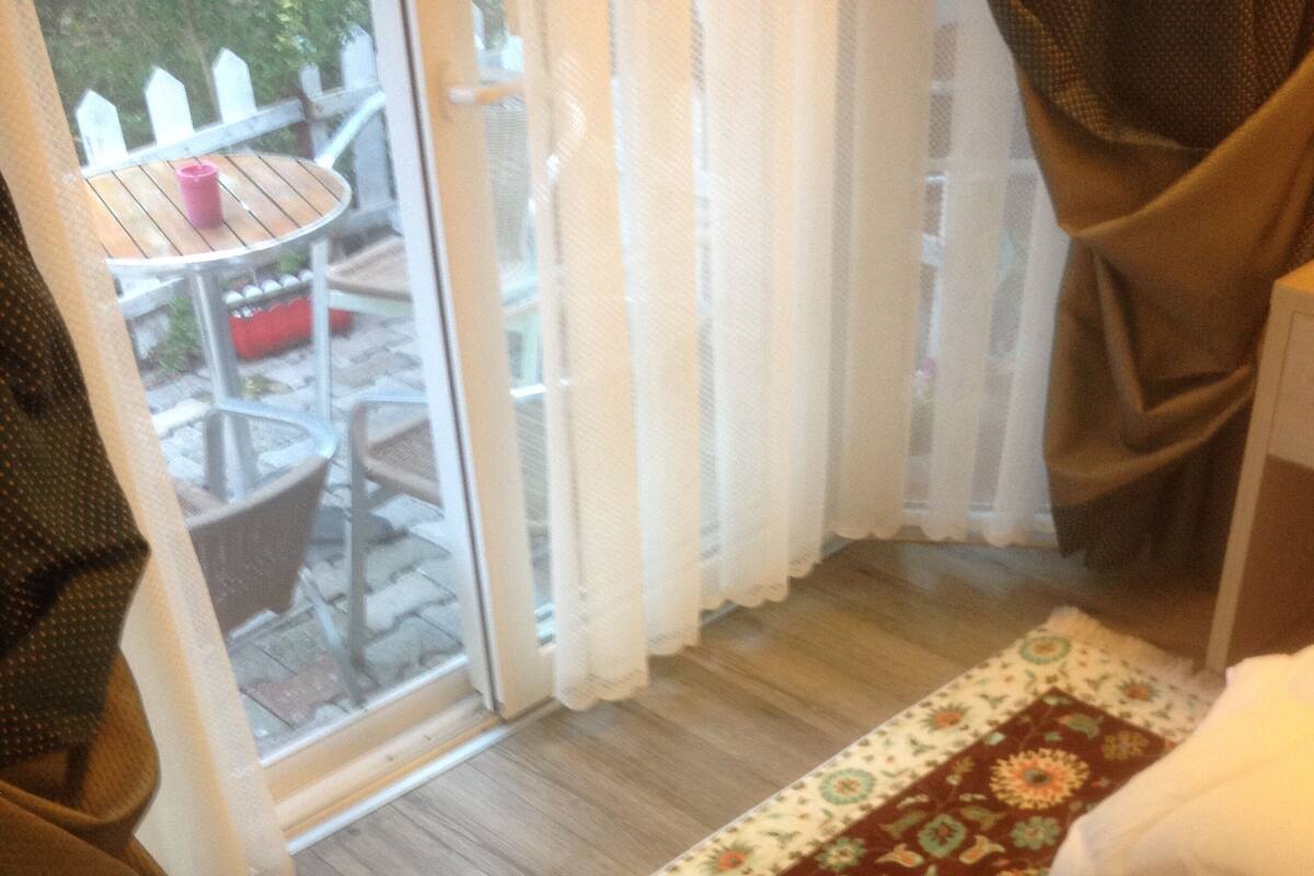 Ein Zimmer mit Holzboden, einem gemusterten Teppich, einem Bett mit weißer Decke, einem Fenster mit Vorhängen und Blick auf eine Terrasse mit Tisch und Stühlen.