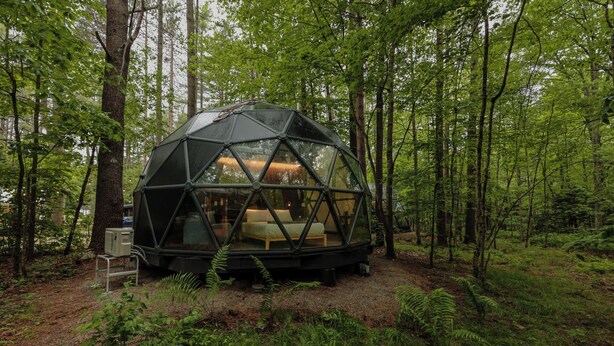 A geodesic dome structure in a forest, with a bed inside and a small table beside it. - 22-ft dome w/ stargazing loft and full bath - 08 (Freeport)