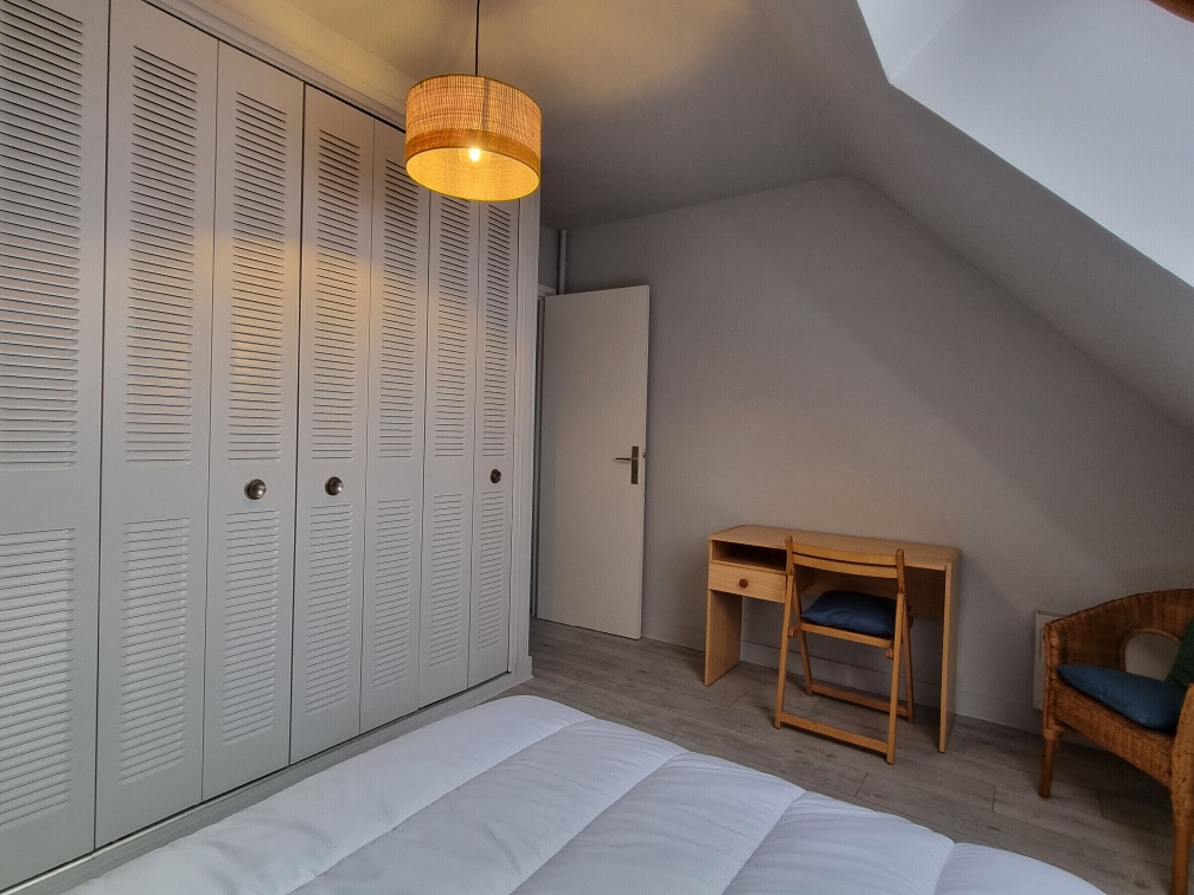 A bedroom with a loft bed, a wooden desk, a wicker chair, and a wardrobe with louvered doors.