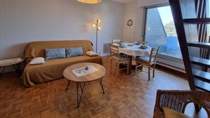 Ein gemütliches Wohnzimmer mit einer Couch, einem Esstisch für vier Personen, einem kleinen runden Tisch mit Untertasse und einer Hängelampe.