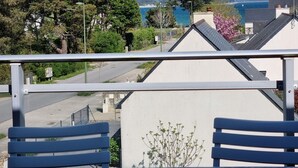 Ein Balkon mit Metallgeländer, der auf einen Wohnbereich mit Häusern, einer Straße und einem Gewässer in der Ferne blickt.