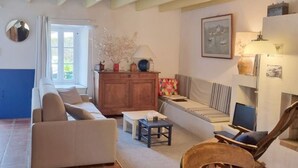 A living room with a sofa, a chair, a coffee table, and a TV. There is a window with curtains, a lamp, and a cabinet with decorative items. - Charming fisherman's house, 4 pers, near beach, Saint-Gildas-de-Rhuys, wifi (Saint-Gildas-de-Rhuys)