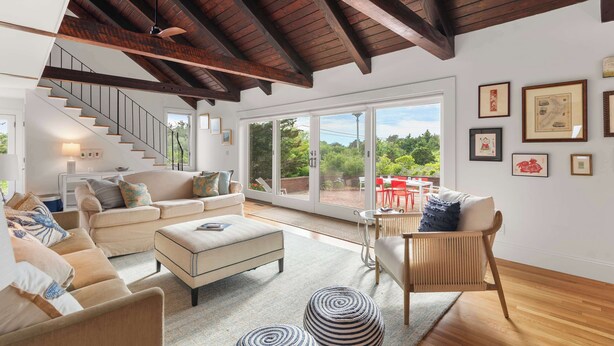 A spacious living area with a high ceiling, wooden beams, a staircase, a large sliding glass door leading to an outdoor patio, and various seating options. - Private Beach, Water Views, Incredible Location! (Chatham)