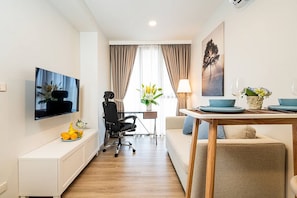 A modern hotel room with a flat-screen TV, a dining table with blue plates, a sofa, a desk with a chair, and a window with curtains.