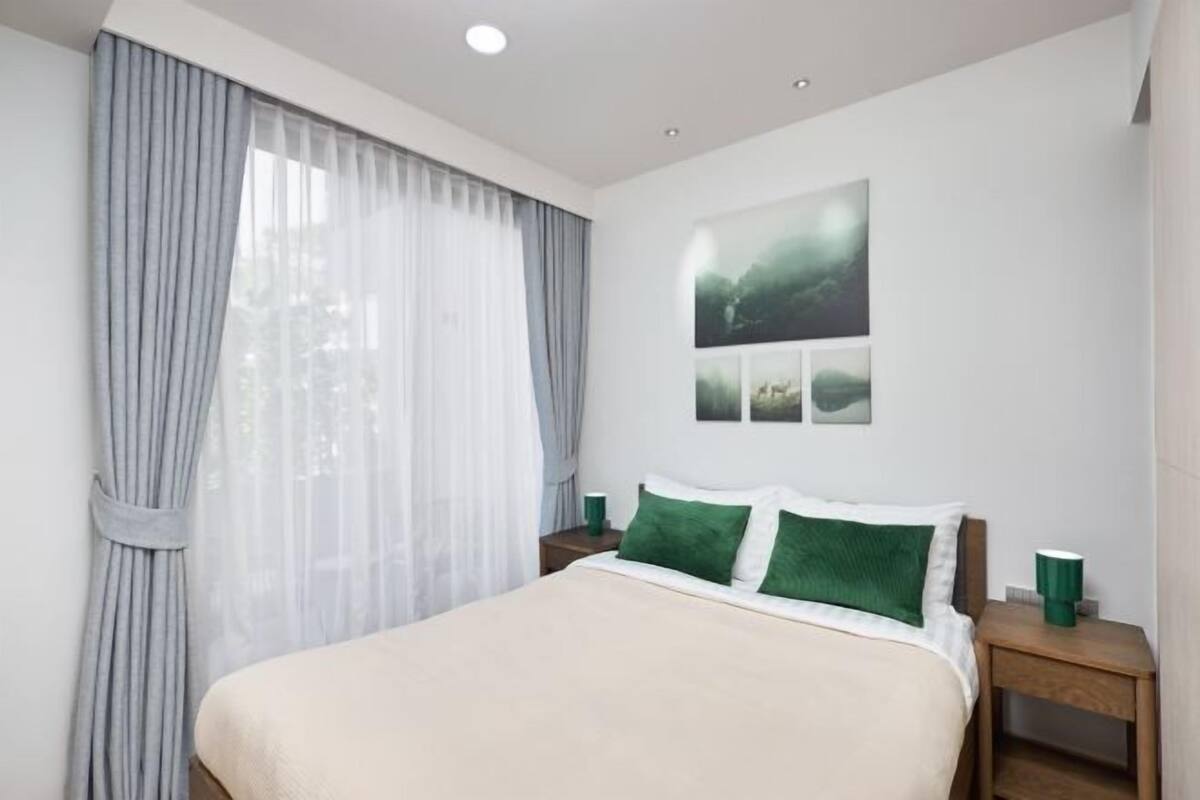Une chambre avec un grand lit, une table de chevet, une tasse verte et une fenêtre ornée de rideaux légers.