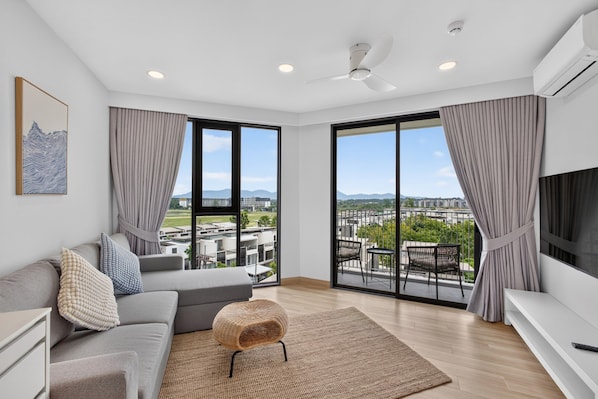 A modern living room with a grey sofa, a woven pouf, and a view of a cityscape through large windows.