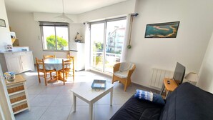 Sala de estar con sofá, una mesa de comedor con sillas, una zona de cocina y ventanas amplias con vistas al exterior.