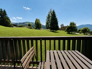 Una terraza de madera con un banco, con vistas a un campo cubierto de hierba, árboles y una montaña al fondo.