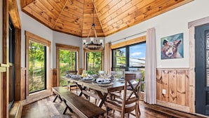 Un coin repas avec une table en bois dressée pour quatre personnes, de grandes fenêtres et un plafond avec des poutres apparentes.